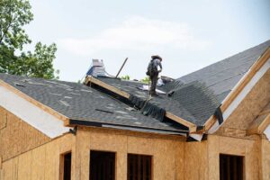 A close-up of a residential roof showing moisture on shingles after rainfall with trees in the background.